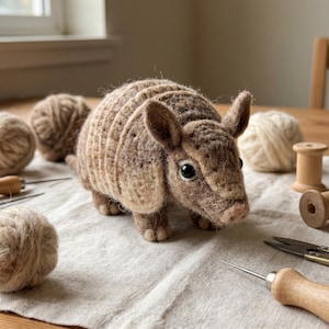May include: A needle-felted armadillo figurine crafted from brown and tan wool fibres. The armadillo features a segmented shell, pointed ears, and dark, glossy eyes. It is positioned among various crafting supplies including yarn balls.
