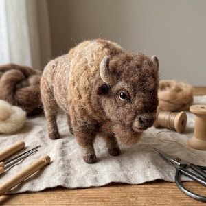 May include: A needle-felted bison figurine crafted from textured brown and tan wool fibers. The sculpture features a thick mane, small curved horns, and dark eyes, standing on a light-colored fabric surface alongside wooden spools and metal scissors.