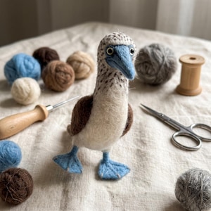 May include: A needle-felted blue-footed booby bird figurine with a textured cream and brown body, a pointed blue beak, and bright blue webbed feet. The bird is surrounded by small balls of wool, a wooden spool, a felting needle, and metal scissors.