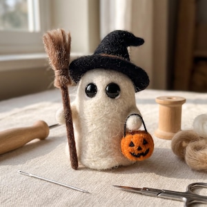 Puede incluir: Una figura de fantasma blanco de lana fieltrada que lleva un sombrero puntiagudo negro con una banda marrón. El fantasma sostiene una escoba marrón en una mano y un pequeño cubo de calabaza naranja en la otra, con grandes ojos negros.