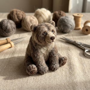 Peut inclure: Une figurine d'ours brun en laine feutrée avec une texture duveteuse, des yeux sombres et un museau clair. L'ours est entouré de plusieurs pelotes de laine, de deux aiguilles à feutrer à manche en bois et d'une paire de ciseaux en métal.