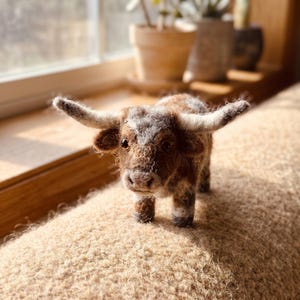 May include: A needle-felted highland cow figurine featuring a textured brown and white coat, long curved horns, and dark eyes. The small animal sculpture stands on four legs and displays a shaggy, woolly appearance with detailed facial features.