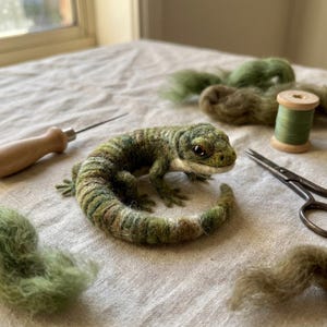 May include: A needle-felted lizard figurine in shades of green and brown with a curled tail and dark eyes. The lizard is surrounded by loose green wool roving, a wooden-handled felting tool, a spool of green thread, and metal scissors.
