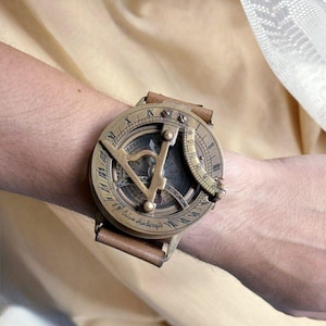 Puede incluir: Un reloj de sol de estilo vintage con esfera de color bronce y correa de cuero marrón. La esfera del reloj presenta números romanos y un diseño de brújula. El texto "Calvin Handicrafts" es visible en el reloj.