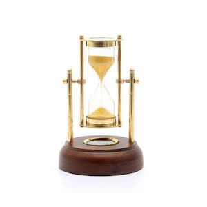 May include: An hourglass with a clear glass bulb containing yellow sand, held within a polished brass frame. The frame is mounted on a circular, dark brown wooden base that features a small, circular brass inlay centered on the top surface.