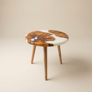 May include: A round side table featuring a natural wood grain top with irregular, organic edges filled with smooth, opaque white resin. The table is supported by three tapered, light brown wooden legs that extend outward from the center.