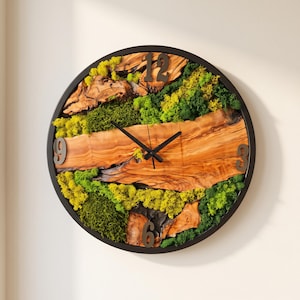 Op de afbeelding: Een ronde wandklok met een wijzerplaat van natuurlijk hout met een levende rand, geaccentueerd door stukken lichtgroen en donkergroen geconserveerd mos. De klok heeft een zwarte rand, zwarte wijzers en grote zwarte cijfers op 12, 3, 6 en 9.