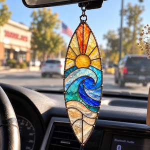 May include: A surfboard-shaped stained glass suncatcher featuring a textured design of a yellow sun rising over blue ocean waves and a sandy beach, held together by dark metal soldering and suspended by a small chain from a rearview mirror.