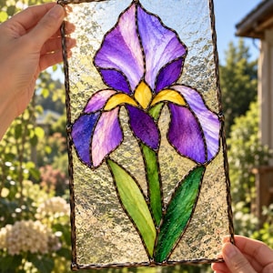 以下が含まれることがあります： 紫と黄色のアイリスの花と緑の葉が描かれた長方形のステンドグラスパネル。質感のある半透明のガラス片が暗い金属のハンダ線で繋ぎ合わされており、鮮やかな花のモチーフが表現されています。