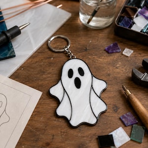 May include: A white stained glass ghost keychain with black glass accents for the eyes and mouth. The ghost figure is outlined in dark metal solder and attached to a circular silver-toned metal split ring for keys, displayed on a wooden surface.
