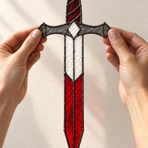 May include: A stained glass sword ornament featuring a circular red pommel, a dark red handle, and a crossguard with a central white diamond. The blade is divided into a white upper section and a vibrant red lower section, all held together by dark metal solder lines.