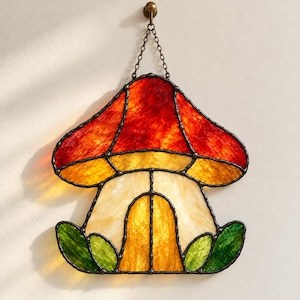 May include: A stained glass mushroom suncatcher featuring a vibrant red and orange cap, a cream-colored stem, and green leaves at the base. The piece is constructed with dark metal soldering and hangs from a delicate metal chain.