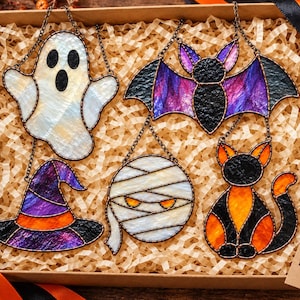 Puede incluir: Un conjunto de cinco adornos de vidrio tintado que incluye un fantasma blanco, un murciélago morado y negro, un sombrero de bruja morado y naranja, una momia blanca y un gato negro y naranja. Cada pieza incluye una cadena de metal para colgar.