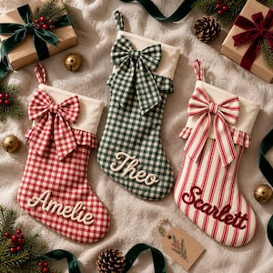 Puede incluir: Tres calcetines de Navidad con puños y lazos de color crema. El calcetín izquierdo presenta un patrón de cuadros vichy rojo y blanco con el nombre Amelie. El central tiene un patrón de cuadros vichy verde y blanco con el nombre Theo. El derecho tiene rayas rojas y blancas con el nombre Scarlett.