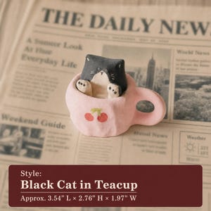 May include: A ceramic figurine of a black and white cat peeking out of a light pink teacup. The teacup features a small red cherry graphic on the front. The dimensions are approximately 3.54 inches long, 2.76 inches high, and 1.97 inches wide.