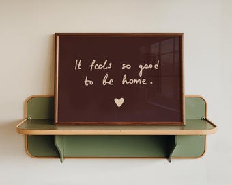 May include: A rectangular dark brown framed print featuring the white text "It feels so good to be home." with a small white heart icon below the text, resting on a green wall-mounted shelf with rounded edges and light-colored trim.