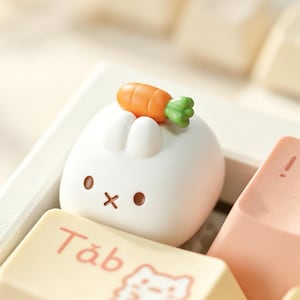 以下が含まれることがあります： 2つの小さな耳、茶色の丸い目、X字型の口を持つウサギの形をした、白く丸みを帯びたメカニカルキーボードのキーキャップ。緑の葉が付いた小さなオレンジ色のニンジンがウサギの頭の上に乗っており、クリーム色のTabキーの隣に配置されています。