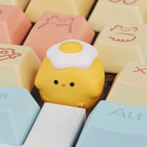 Peut inclure: Une touche de clavier mécanique en forme de poussin jaune avec un œuf au plat blanc sur la tête, placée parmi d'autres touches de couleur claire avec divers dessins au trait et le texte Alt imprimé sur l'une des touches bleues environnantes.