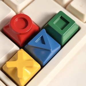 May include: Four custom mechanical keyboard keycaps featuring raised geometric symbols. A red circle, a green square, a blue triangle, and a yellow cross are positioned on a white keyboard base, replacing standard keys with distinct shapes and colors.