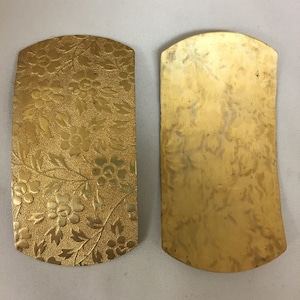 May include: Two gold-colored metal belt buckles. One buckle has a floral pattern, while the other is plain.