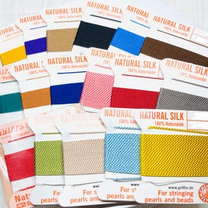 May include: A collection of Griffin Natural Silk cord spools in various colors including yellow, purple, tan, black, blue, brown, teal, orange, pink, red, grey, lime green, and light blue, each labeled Natural Silk 100% Naturseide for stringing beads.