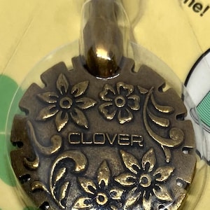 May include: A gold-toned metal thread cutter with a floral design and the word "CLOVER" inscribed on it. The cutter has a loop at the top for hanging.
