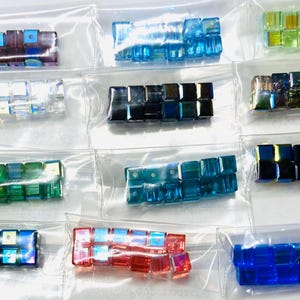 May include: A collection of colorful, square glass beads in small, clear plastic bags. The beads are various colors, including blue, green, purple, and iridescent. The beads are likely for jewelry making or other crafts.