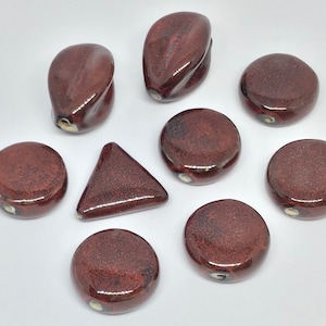 May include: A collection of nine ceramic beads in various shapes, including round, oval, and triangular. The beads are a deep reddish-brown color with a glossy finish.