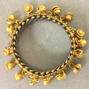 May include: A gold-toned charm bracelet with a silver-colored, expandable band. The bracelet features various small, gold-colored charms, including bell-shaped and other decorative elements. The bracelet is designed to fit a range of wrist sizes.