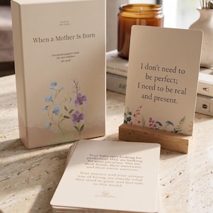 May include: A deck of 40 emotional support cards for new mothers titled When a Mother Is Born, featuring a beige box with floral illustrations, a wooden card stand, and a card displaying the text I don't need to be perfect; I need to be real and present.