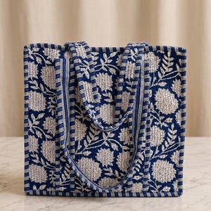 May include: A square quilted tote bag featuring a deep blue background with a white floral pattern. The bag has two matching fabric handles and is accented with vertical striped trim along the edges and the straps, creating a structured look.
