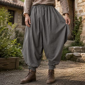 Op de afbeelding: Losvallende antracietgrijze broek met een geplooide elastische tailleband met trekkoord, een verlaagd kruis en taps toelopende boorden bij de enkels. De stof heeft een matte textuur en valt in zachte, verticale plooien.