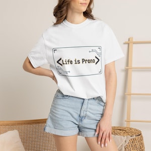 Può includere: T-shirt bianco con la scritta "Life is Prono" all'interno di un design rettangolare con dettagli in stile circuito stampato. La maglietta è a girocollo e si abbina a pantaloncini di jeans. L'aspetto è casual e moderno.