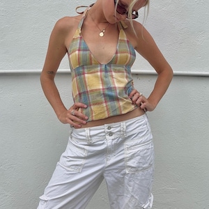 May include: A halter top featuring a yellow, blue, and red plaid pattern with a deep V-neckline and tie-neck closure, paired with white cargo pants that have a double-button waistband and multiple utility pockets on the front.