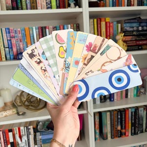 May include: A collection of rectangular bookmarks fanned out in a hand, featuring diverse designs including a blue evil eye pattern, floral motifs, fruit illustrations like watermelon and avocado, green gingham, and various colorful graphic prints.