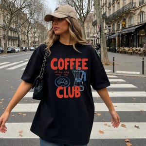 May include: Black t-shirt with the words "COFFEE CLUB" in red and a blue coffee pot graphic. The shirt also features the text "all day every day" and a croissant illustration. A beige baseball cap and a black purse complete the look.