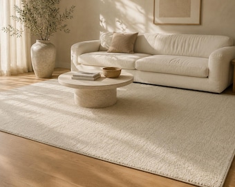 May include: A large, rectangular area rug in a solid cream color with a textured, low-pile surface. The rug is positioned on a light-toned wooden floor, covering a significant portion of the space in front of a sofa and a round stone coffee table.
