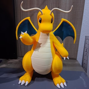Puede incluir: Una figura de dragón amarilla con vientre color crema, alas azul oscuro y garras blancas. La criatura tiene dos antenas largas y curvas en la cabeza y ojos grandes y oscuros. A la derecha se ve una regla metálica con medidas en centímetros.