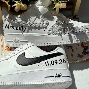 May include: Two white sneakers with custom details. The top shoe features white pearls and clear crystals along the side, with the text Mrs. Lilly printed in black. The bottom shoe has a black swoosh logo and the date 11.09.26 printed in black.