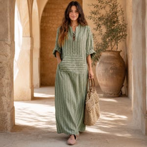 May include: A long-sleeved, floor-length dress in a muted sage green with vertical white stripes. The dress features a split neckline, a large front kangaroo pocket, and loose-fitting sleeves. A woven tan net tote bag with circular handles is carried.