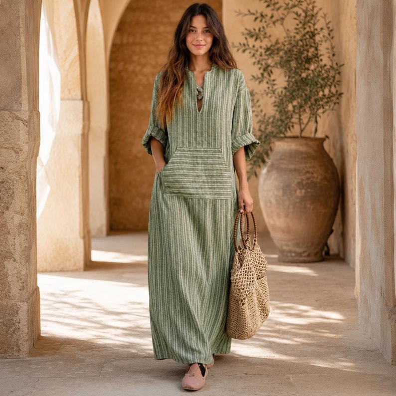 May include: A long-sleeved, floor-length dress in a muted sage green with vertical white stripes. The dress features a split neckline, a large front kangaroo pocket, and loose-fitting sleeves. A woven tan net tote bag with circular handles is carried.