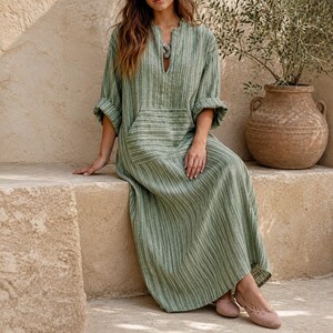 May include: A long-sleeved, floor-length dress in a sage green and white vertical striped pattern. The dress features a split V-neckline, a large front patch pocket, and loose-fitting sleeves with rolled cuffs, creating a relaxed, flowing silhouette.