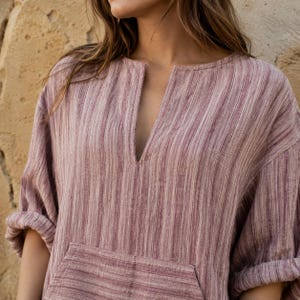 May include: A mauve and white striped tunic featuring a deep vertical slit neckline, long sleeves with rolled cuffs, and a large front kangaroo pocket. The garment is crafted from a textured fabric with a subtle vertical pattern throughout.