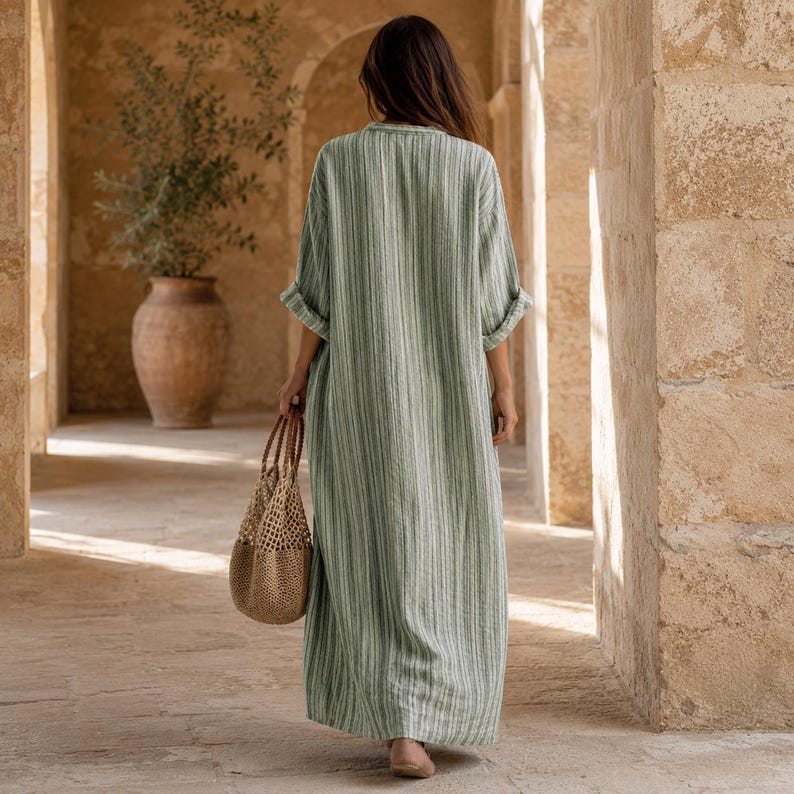 May include: A floor-length, sage green and white striped dress with three-quarter length sleeves and a loose, flowing silhouette. The garment is paired with a tan, woven net-style tote bag featuring a circular base and a single handle.