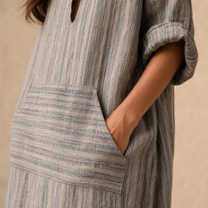 May include: A long-sleeved tunic dress featuring a vertical blue and beige striped pattern. The garment includes a large front kangaroo pocket and a notched neckline. The sleeves are rolled up at the elbows, and the fabric has a textured, woven appearance.