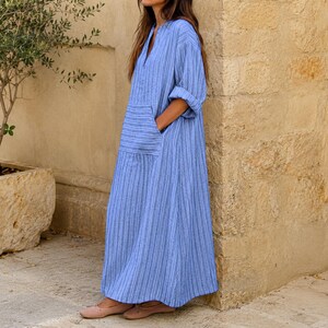 May include: A long, loose-fitting blue dress featuring a vertical white pinstripe pattern, a V-neckline, three-quarter length sleeves with rolled cuffs, and a large patch pocket on the front. The garment reaches the ankles and has a relaxed silhouette.