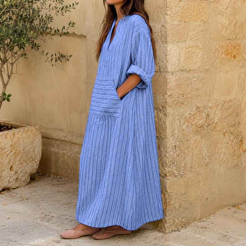 May include: A long, loose-fitting blue dress featuring a vertical white pinstripe pattern, a V-neckline, three-quarter length sleeves with rolled cuffs, and a large patch pocket on the front. The garment reaches the ankles and has a relaxed silhouette.