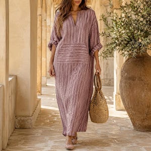 May include: A long-sleeved, floor-length dress in a mauve and white vertical striped pattern. The dress features a V-neckline, a large rectangular front pocket, and loose-fitting sleeves. A woven, tan-colored straw handbag with two handles is held.