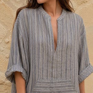 May include: A loose-fitting tunic featuring a vertical striped pattern in shades of grey, blue, and beige. The garment has a split V-neckline, three-quarter length sleeves with folded cuffs, and a large rectangular front pocket with horizontal stripes.