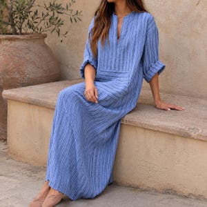 May include: A long-sleeved maxi dress in a blue and white vertical striped pattern. The dress features a split V-neckline, a relaxed fit, and a large horizontal front pocket. The sleeves are cuffed at the elbows, and the fabric has a textured appearance.